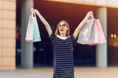 Heyecanlı, şatafatlı, güneş gözlüklü, renkli alışveriş çantaları olan alışveriş merkezinin arka planında caddede duran mutlu, şık bir kadın. Alışveriş, moda.