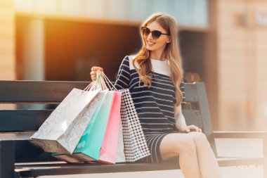 Güneş gözlüklü çekici genç bir kadın alışveriş merkezinin yanındaki bankta oturuyor. Alışveriş, moda.
