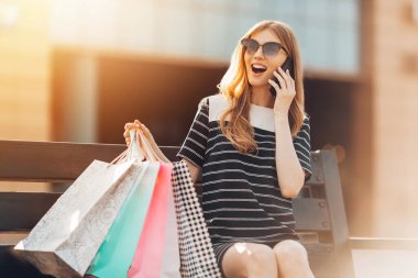 Güneş gözlüğü takan mutlu genç bir kadın alışveriş merkezinin arkasındaki bankta oturuyor ve cep telefonuyla konuşuyor. Alışveriş, moda.