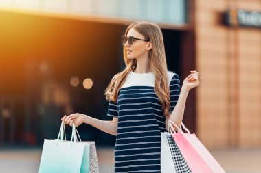 Güneş gözlüklü, çok renkli alışveriş çantalı mutlu ve güzel bir kadın alışveriş merkezinin önünde sokakta alışveriş yapıyor. Alışveriş, moda.
