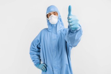 Koronavirüse karşı koruyucu kıyafet giyen, tıbbi maske takan, gözlük takan ve eldiven takan erkek doktor, izole edilmiş beyaz bir arkaplanı onaylıyor. Karantina, koronavirüs