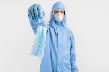 Koronavirüse karşı koruyucu giysi giyen, tıbbi maske, gözlük ve eldiven takan erkek doktor, beyaz arka planda koruyucu bir maske tutuyor. Karantina, koronavirüs