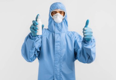 Koronavirüse karşı koruyucu kıyafetli bir erkek doktor, eldivenler, gözlük ve tıbbi maske takıyor, izole edilmiş beyaz bir arka planda bir kavanoz antiseptik tutuyor. Karantina, koronavirüs