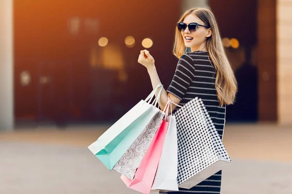 在一个购物中心的背景下 一个戴着太阳镜 手里拿着购物袋 面带微笑的时髦女孩 — 图库照片