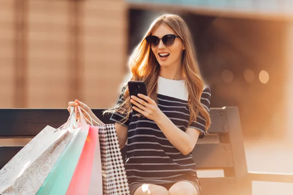 attractive happy beautiful young woman in sunglasses is sitting on a bench near a shopping center with shopping bags, using a mobile phone. Shopping, fashion