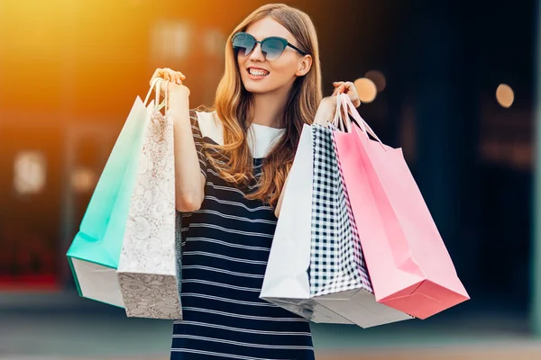 一个快乐 戴着太阳镜 头戴五颜六色购物袋的年轻女子正在购物中心前的街上购物 — 图库照片