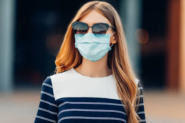 サングラスの若い魅力的な女の子と彼女の顔の医療保護マスク建物の背景に街に立っている 隔離コロナウイルス — ストック写真