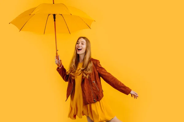Happy girl in a yellow dress and autumn jacket, laughing and having fun with a yellow umbrella, standing on a yellow background