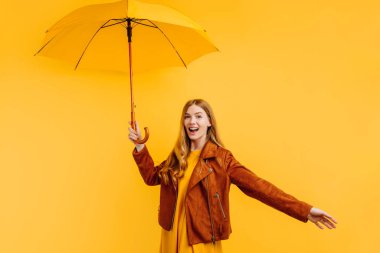 Sarı elbiseli ve sonbahar ceketli mutlu duygusal kız, sarı arka planda sarı bir şemsiyeyle gülüyor ve eğleniyor.