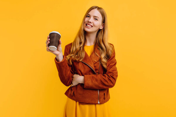 Stylish happy beautiful young woman in a yellow dress and autumn jacket, holding a paper Cup of coffee, on an isolated yellow background. The concept of autumn