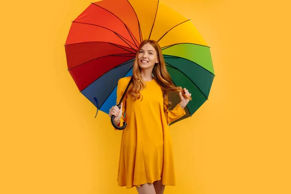 Happy attractive girl, in a yellow dress, happy and posing with a rainbow umbrella, on an yellow background