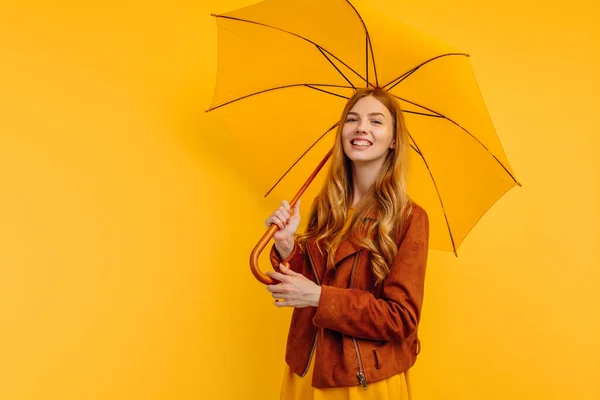 happy attractive girl, in a bright yellow dress and autumn jacket, stands with a yellow umbrella on an isolated yellow background