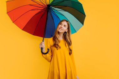Sarı elbiseli mutlu kız, sarı bir arka planda gökkuşağı şemsiyesiyle düşünüyor ve poz veriyor.