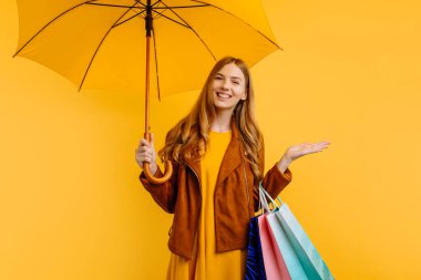 Gülümseyen kız, parlak sarı bir elbise ve sonbahar ceketi içinde, sarı bir şemsiye ve alışveriş çantasıyla duruyor, sarı bir arka planda.