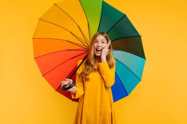 Şok olmuş şaşırmış, sarı elbiseli, mutlu ve izole edilmiş sarı bir arka planda gökkuşağı şemsiyesiyle poz veren bir kız.