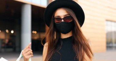 Yüzünde tıbbi bir maskeyle, siyah şık bir elbise ve şapka giymiş, alışveriş torbalarıyla alışveriş yaparken, Kara Cuma, alışveriş yaparken mutlu, modaya uygun genç bir kadın.