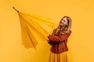 Sarı elbiseli ve sonbahar ceketli mutlu çekici kız izole edilmiş sarı arka planda sarı bir şemsiye açıyor.