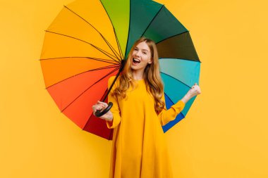 Şok olmuş mutlu kız, sarı elbiseli, mutlu ve gökkuşağı şemsiyesiyle poz veriyor, sarı arka planda kazanan bir jest gösteriyor.