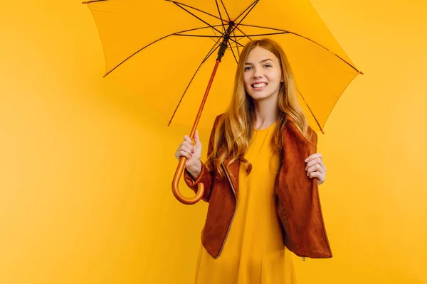 happy attractive girl, in a bright yellow dress and autumn jacket, stands with a yellow umbrella on an isolated yellow background. concept of autumn