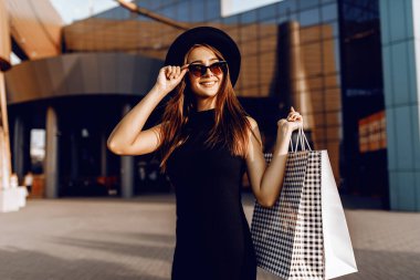 Siyah elbiseli ve şapkalı çekici bir kız, siyah gözlükler takıyor, çantalarını tutuyor ve alışverişin tadını çıkarıyor, Kara Cuma konsepti, alışveriş