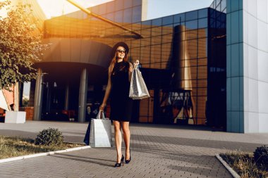 Siyah elbiseli, şapkalı ve güneş gözlüklü şık bir kız alışveriş merkezinin arka planında alışveriş torbalarıyla ayakta duruyor. Alışveriş, Kara Cuma
