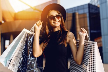 Siyah elbiseli ve şapkalı, siyah gözlükler takan, çanta taşıyan ve alışveriş yapmaktan zevk alan mutlu bir kız.