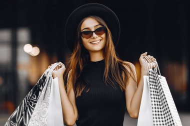 Siyah elbiseli, şapkalı, siyah gözlüklü, alışveriş torbası tutan, alışveriş merkezinin arka planında başarılı bir alışveriş yapan mutlu ve çekici bir genç kadın. Kara Cuma, alışveriş.