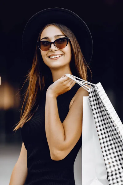 一个漂亮的年轻女人 穿着黑色的衣服 戴着黑色的帽子 戴着深色的眼镜 提着购物袋 站在购物中心的背景下 黑色星期五 — 图库照片