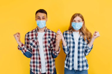 Yüzlerinde tıbbi koruyucu maskeler olan mutlu bir adam ve kadın, ekose gömlekler giyip, eğleniyorlar ve izole edilmiş sarı bir arka planda durup zafer jesti gösteriyorlar..