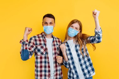 Yüzlerinde tıbbi koruyucu maskeler olan mutlu bir adam ve kadın, ekose gömlekler giyip, eğleniyorlar ve izole edilmiş sarı bir arka planda durup zafer jesti gösteriyorlar..