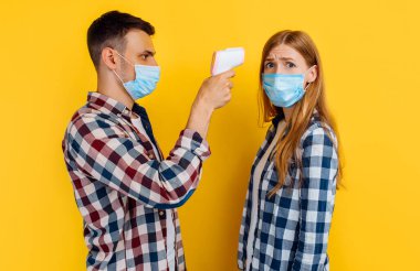 Koronavirüsten dolayı yüzünde tıbbi koruyucu maske olan bir adam ve bir kadın sıcaklığı dijital termometreyle kontrol ediyor ve sarı arka planda duruyor.