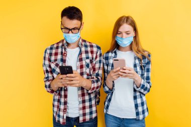 Genç bir çift, bir erkek ve bir kız gömlekli ve koruyucu maskeli, sarı arka planda izole edilmiş cep telefonları kullanıyorlar.