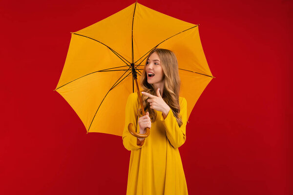 happy young woman with a yellow umbrella pointing at a copy of the space on an empty red background, autumn concept