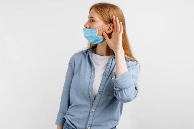 Yüzünde tıbbi bir maske olan meraklı, odaklanmış genç bir kadın, elini kulağına dayayıp, beyaz bir arka plana doğru dinliyor.
