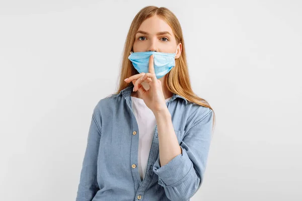 young woman in a medical protective mask on her face, pressing a finger to her lips, makes a gesture of silence, speaks in a low voice, against a white background