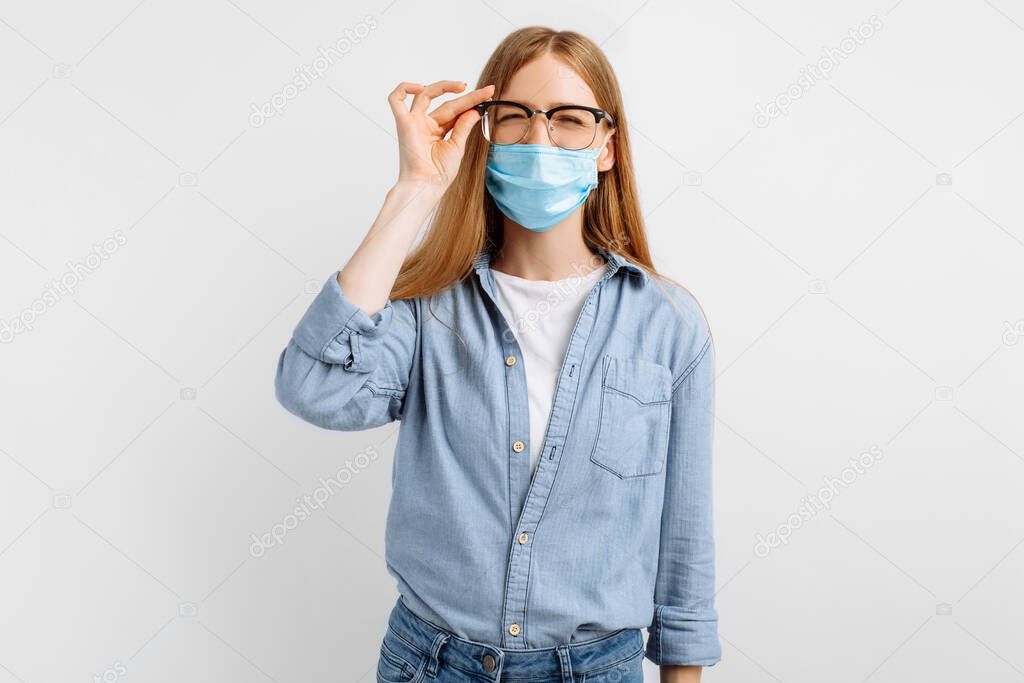 young woman in a medical protective mask on her face, wearing glasses for vision squints at the camera, having vision problems, on a white background