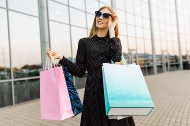Güneş gözlüğü takan ve alışveriş merkezinin arka planında alışveriş çantaları olan mutlu ve güzel bir kadın.