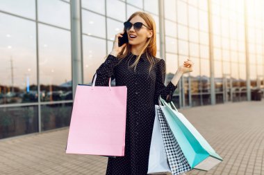 Güneş gözlüklü, alışveriş çantalı, bir alışveriş merkezinin arka planında cep telefonuyla konuşan mutlu genç bir kadın.