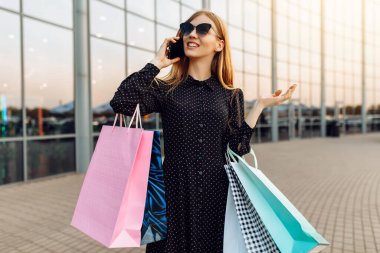 Güneş gözlüklü, alışveriş çantalı, cep telefonuyla konuşan mutlu genç bir kadın.