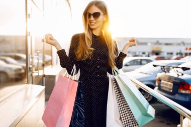 Güneş gözlüğü takan ve alışveriş merkezinin arka planında alışveriş torbaları olan mutlu genç bir kadın.