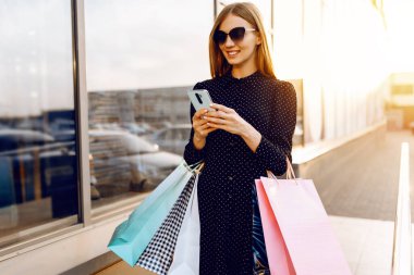 Güneş gözlüklü, alışveriş çantalı mutlu genç bir kadın, bir cep telefonu kullanıyor, bir alışveriş merkezinin arka planında duruyor.