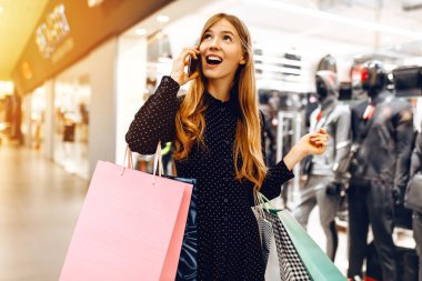 Alışveriş torbalarıyla, alışveriş merkezinde cep telefonuyla konuşan güzel genç bir kadın.