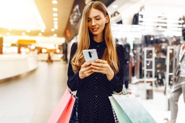 Elbise giymiş güzel genç bir kadın, alışveriş merkezinde cep telefonu kullanıyor.