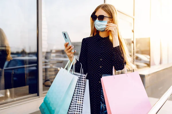 时尚的年轻女性戴着太阳镜 脸上戴着医疗保护面具 头戴购物袋 在市中心的一个购物中心里用手机购物 — 图库照片