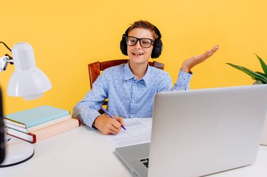 Çocuklar için online dersler. Çocuklar için evde eğitim ve uzak eğitim. Okul çocuğu masada oturuyor, öğretmeniyle video bağlantısı kurarak öğreniyor, sarı arka planda.