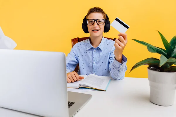 男生拿着笔记本电脑坐在桌旁 手里拿着一张黄色背景的信用卡 网上购物 眼镜蛇 — 图库照片