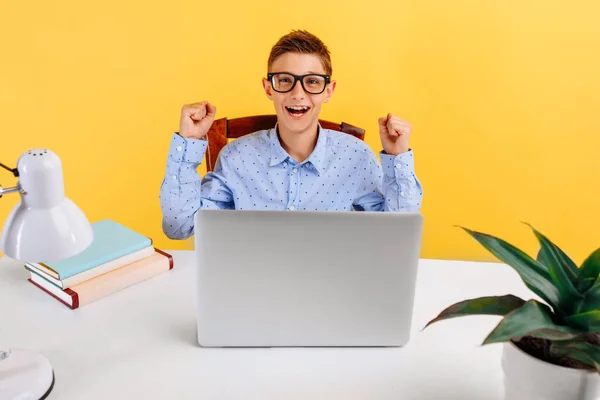 schoolboy sitting at a table, learning by video link, happy and shows a winning gesture on a yellow background. Online lessons for children. distance education for children.