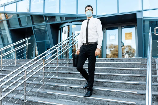 young businessman in business clothes, a man in a medical protective mask on his face, leaves the office building after a working day