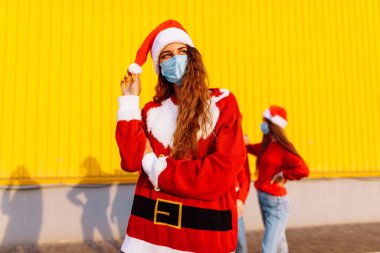 Dört genç arkadaş, Noel süveteri ve Noel Baba şapkaları takan dört genç kadın ve yüzlerinde tıbbi koruyucu maskeler, dışarıda sarı bir duvarın önünde duruyorlar, Noel, koronavirüs.