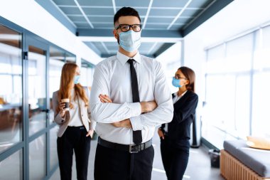Kendine güvenen genç iş adamı lideri yüzünde tıbbi koruyucu bir maske takıyor ve arka planda bir grup meslektaşı bir ofis binasında duruyor.